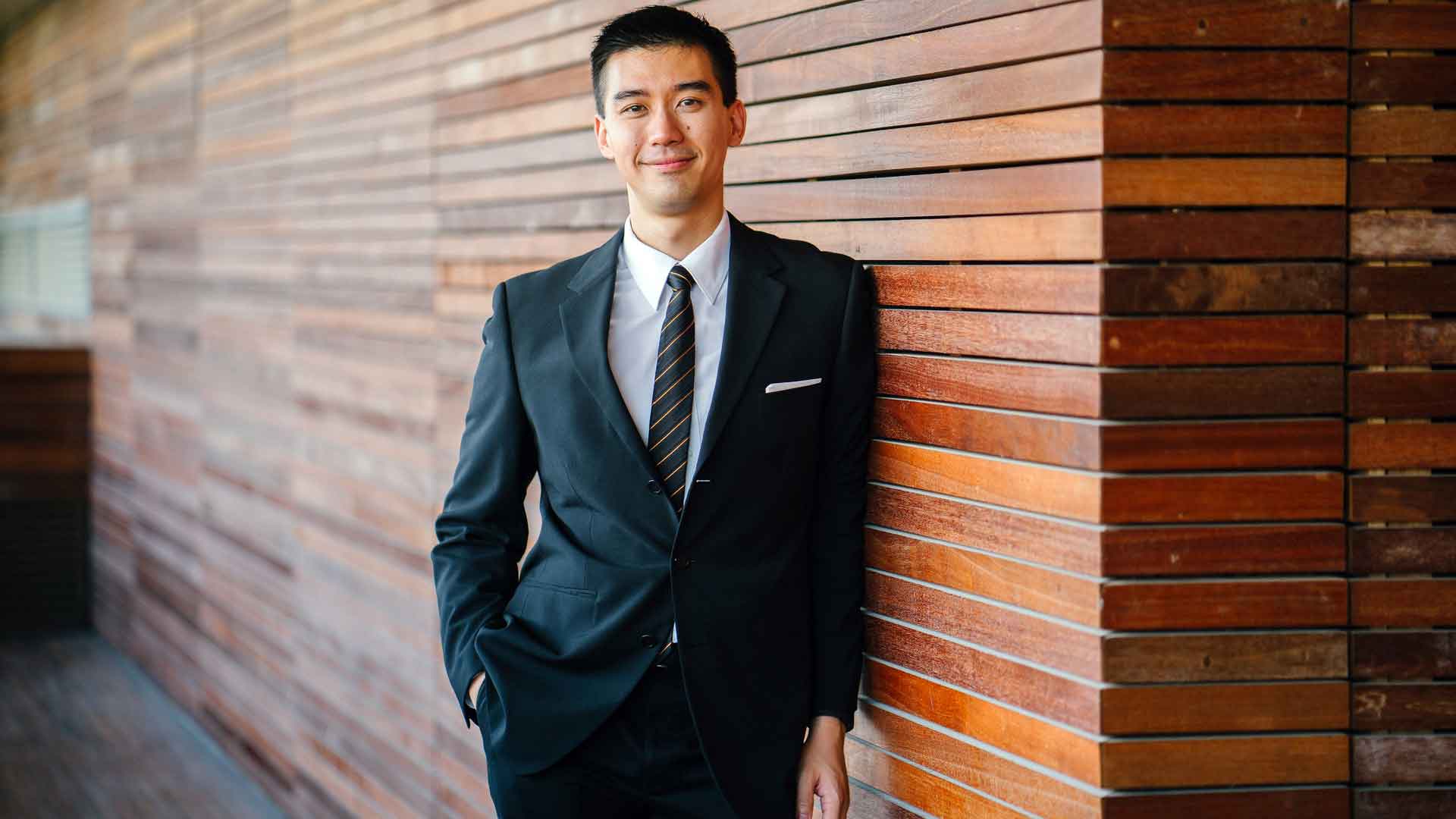 A man smiling wearing suit and tie, standing beside a wooding wall.