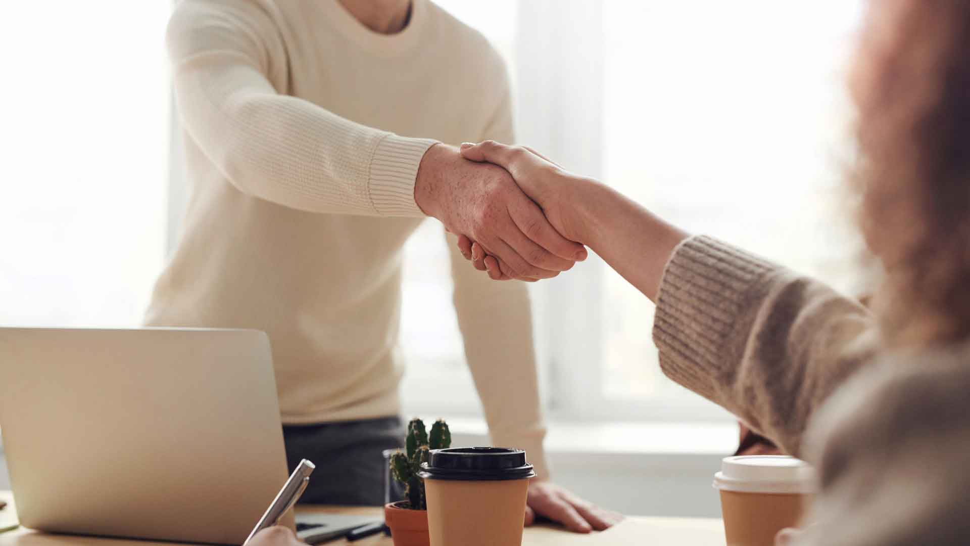 Two person shaking hands in an office with a laptop, cactus plant, and two cups on the table.